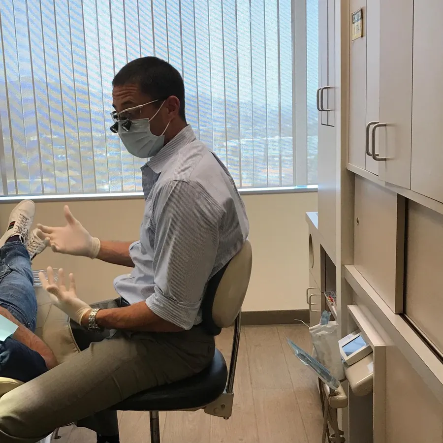 A dentist in a mask and loupes talking with a patient in a treatment room, the window behind him.